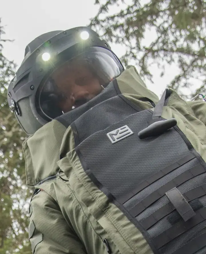 Bomb disposal technician in a Med-Eng EOD bomb suit and helmet with lights on, seen from below next to a vehicle